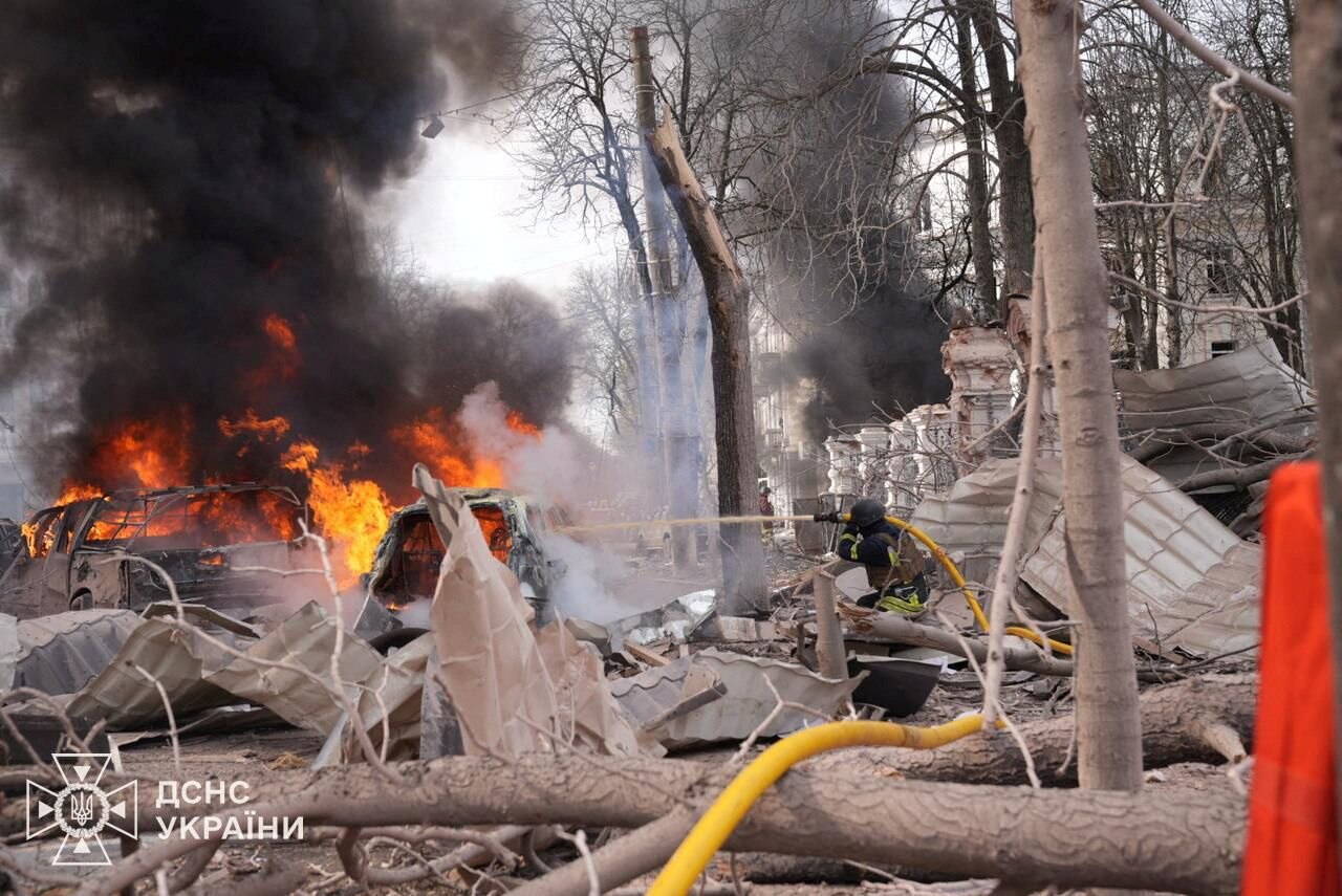 A firefighter using a hose hoisted on their shoulder and aimed at orange flames erupting from a burnt-out car