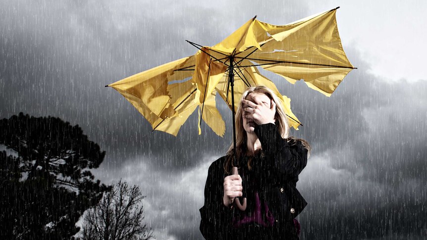 A blonde woman covers her face and stands under a ripped yellow umbrella.