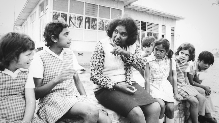 An Indigenous woman sitting with a group of six young children in front of a house, in the 1970s