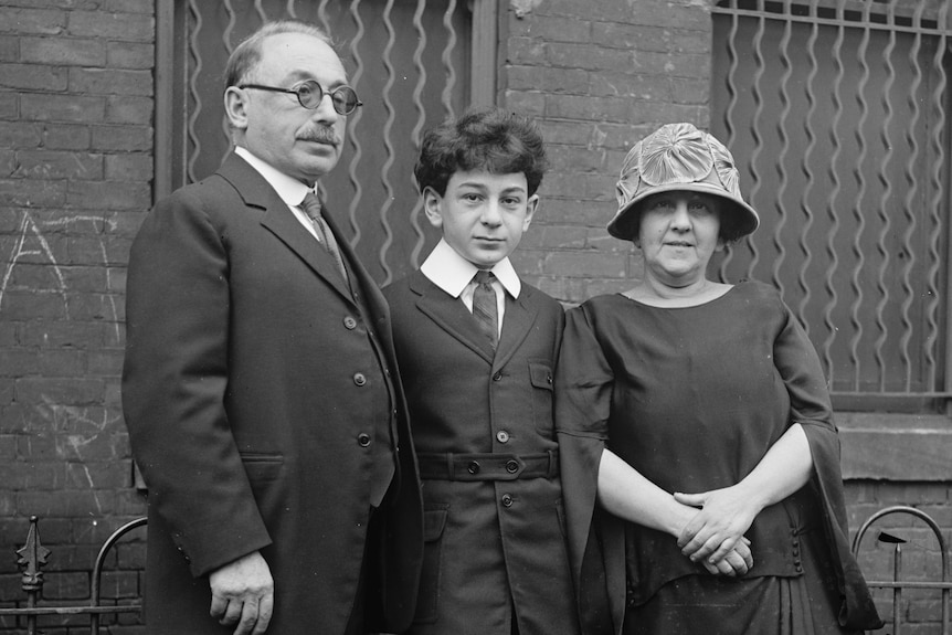 A teenage boy stands between his father and mother in front of a building. They are in formal attire.