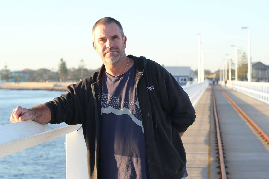 Jon Eddy, un homme de 50 ans portant un t-shirt noir et une veste polaire, debout sur une longue jetée en bois.  C'est tôt le matin.