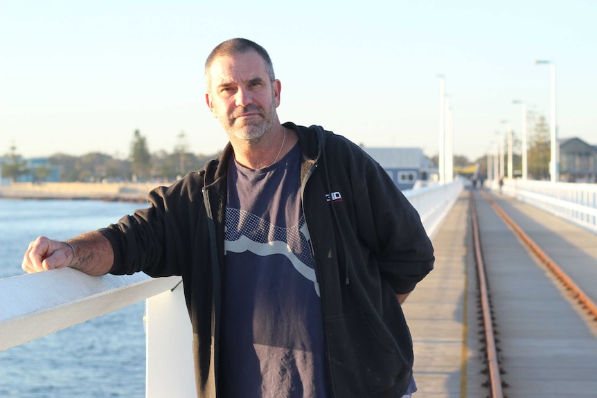 Jon Eddy, A 50-year-old man in a black t-shirt and fleece jacket, standing on a long timber jetty. It's early morning.