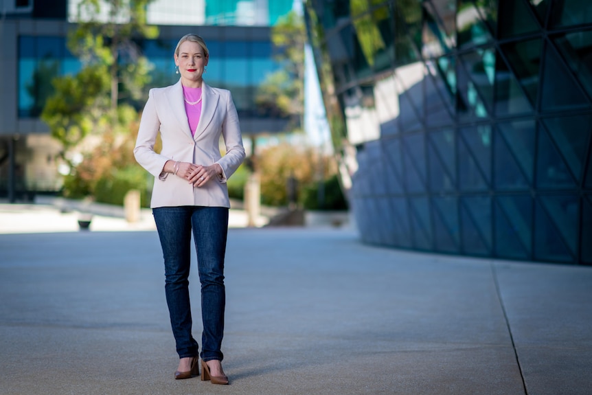 A woman stands next to a hospital building with her hands held at her waist and a serious expression