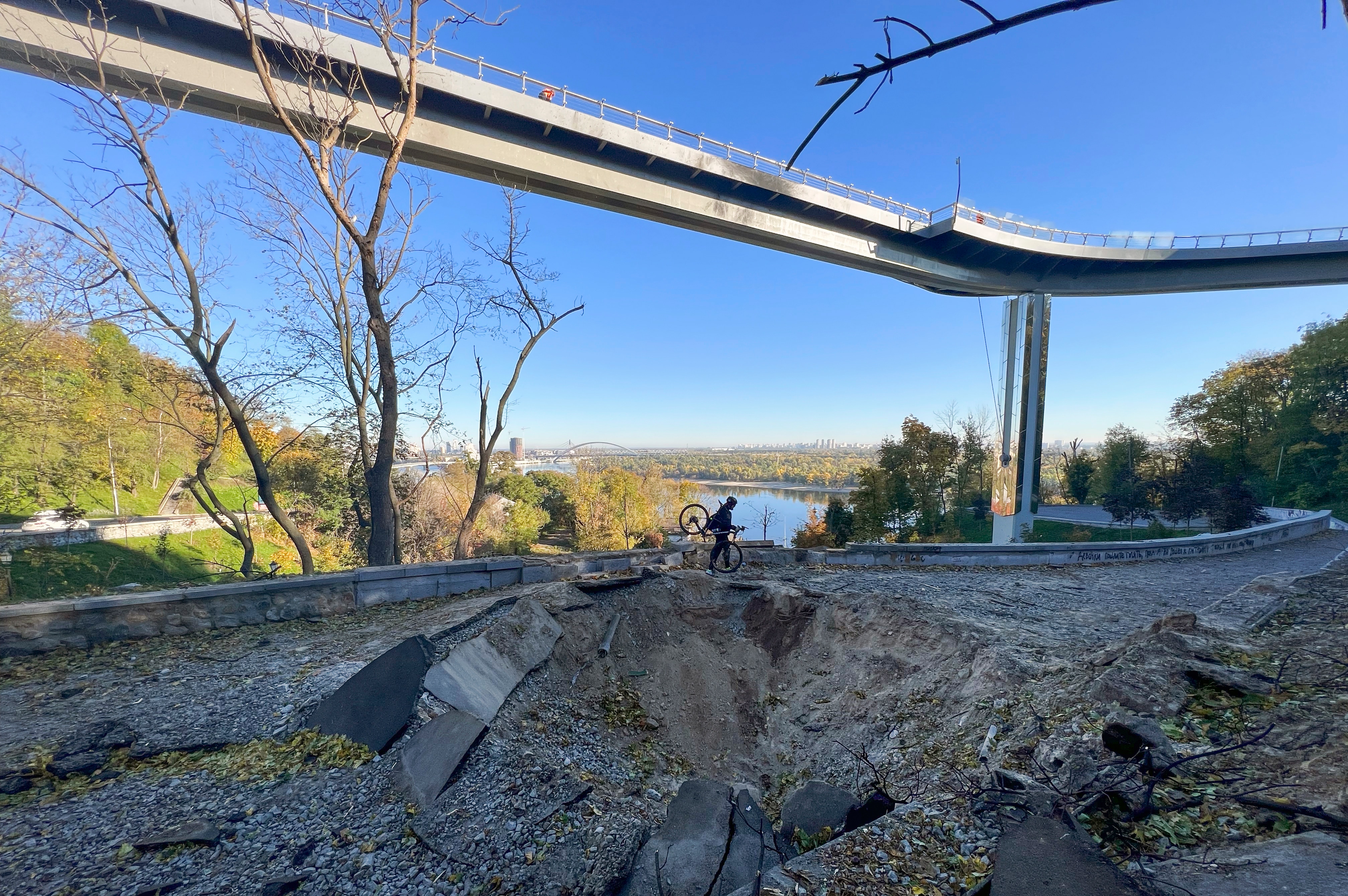 A man carries his bike past a rocket crater under a pedestrian bridge.