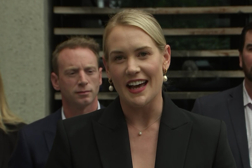 A woman with blonde hair smiles at the camera standing in front of two men