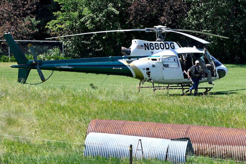 A man with a gun emerges from a helicopter which landed in a field.