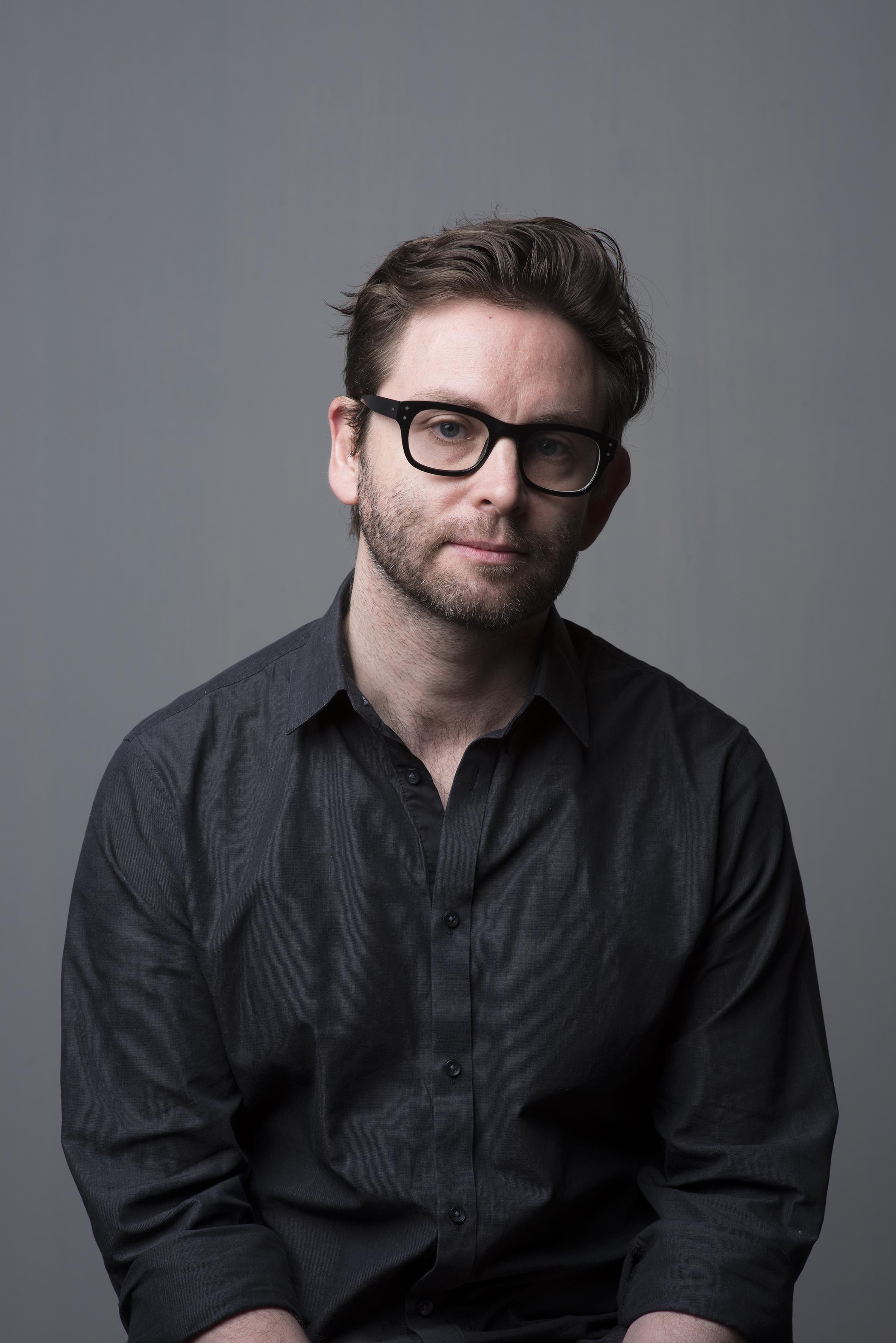 A 40-something man with thick black glasses looks into the camera. He has a beard and is wearing a black button-up shirt.