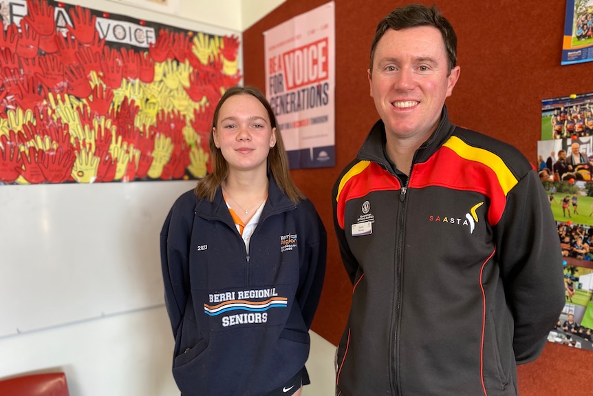 A young woman and man stand smiling. She wears a navy school jumper, he wears black, red and yellow jumper, 