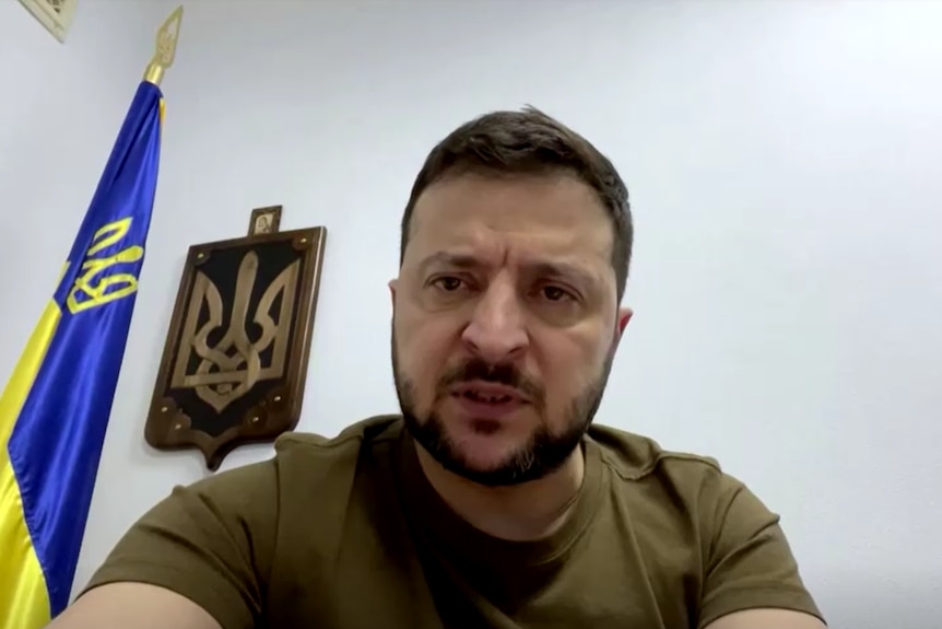 Man in army t-shirt speaks to camera with Ukrainian flag in background.