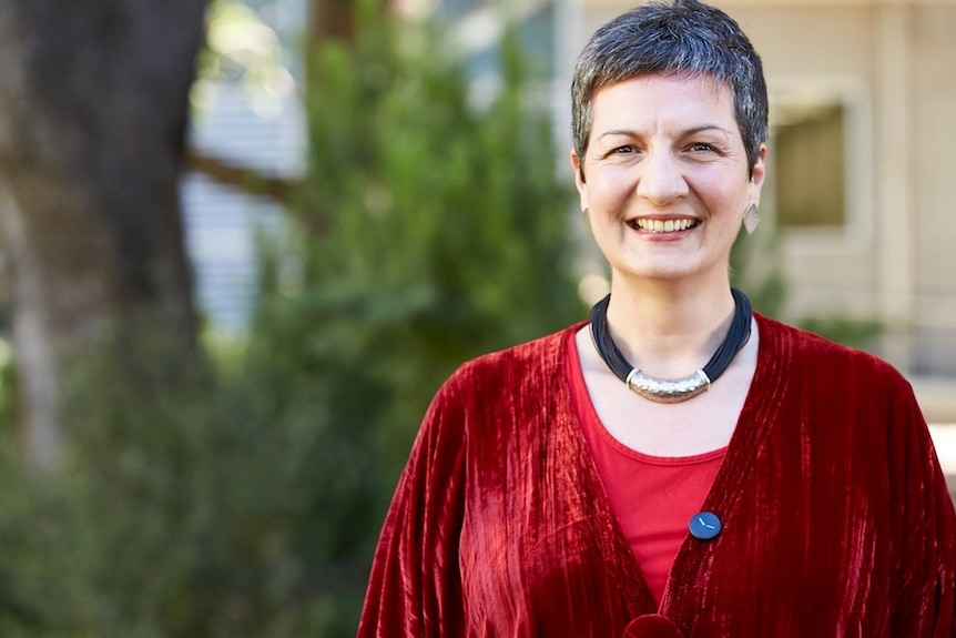 A woman with short hair stands smiling in front of a tree.