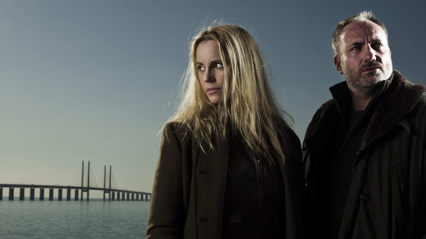 A man and a woman stand against a moody background with a bridge