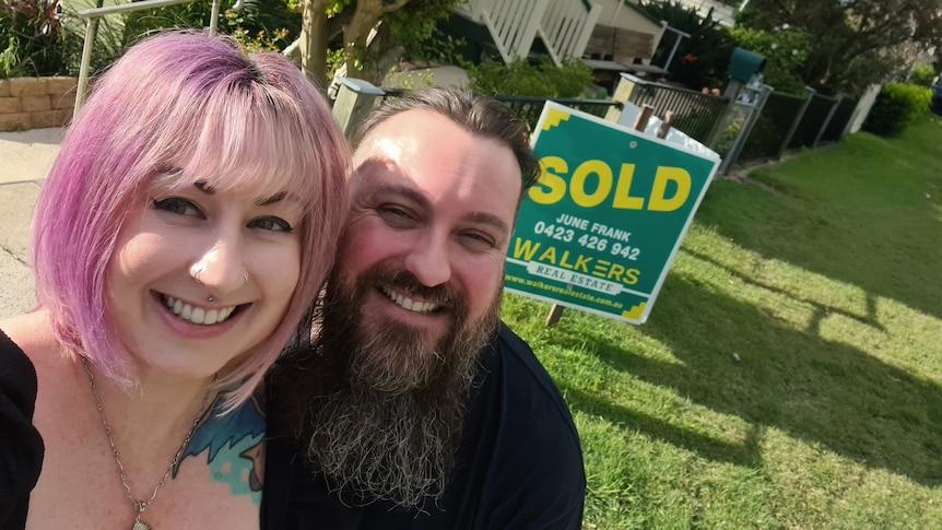 Nick Rusis and Chloe Murdoch smiling in the foreground outside their new Ipswich home with a SOLD sign out the front