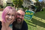 Nick Rusis and Chloe Murdoch smiling in the foreground outside their new Ipswich home with a SOLD sign out the front