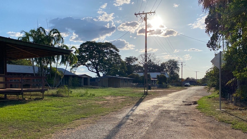 A rough-looking gravel road lined by green grass and a couple of palm trees, with a sunset and telegraph pole in the distance.
