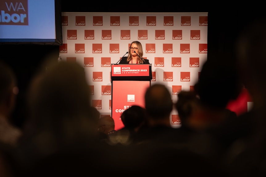 A woman with long blonde hair speaks from a lectern against a white and red background, seen through a crowd of people.
