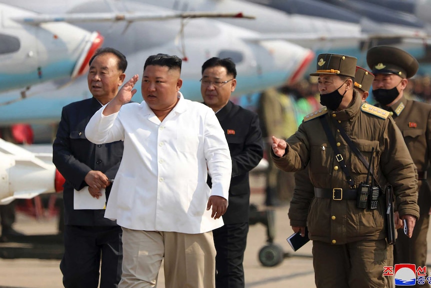 Kim Jogn-un wears a white top and is flanked by military men as they walk past jet planes.