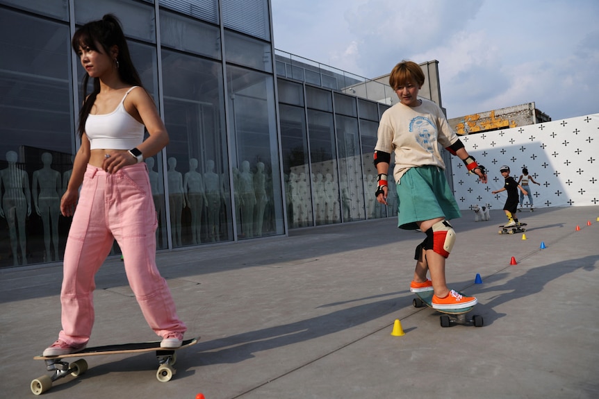 Two women ride skateboards through cones on cement. 