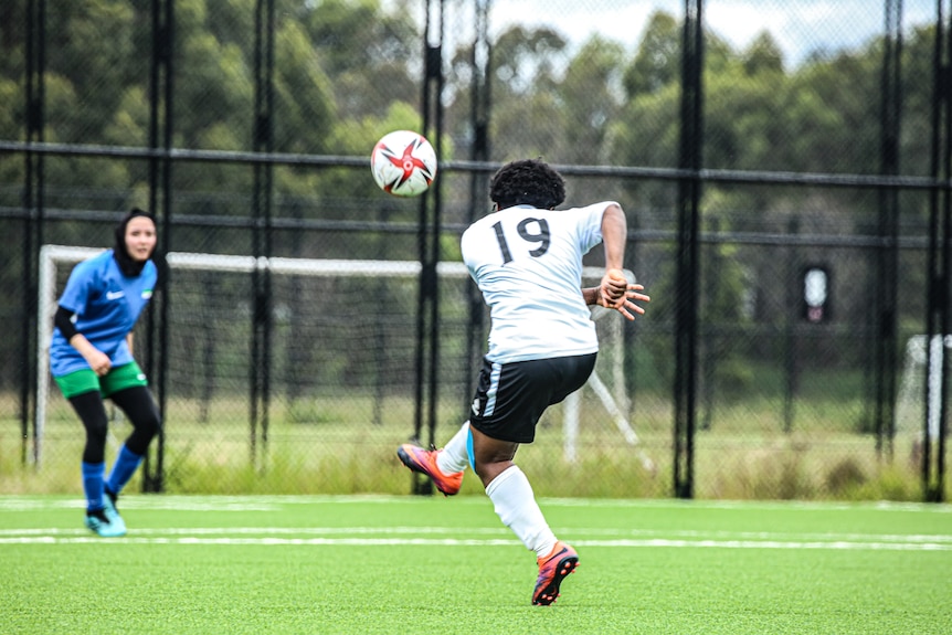 A woman with her back to camera, kicks a football and a defender stands in front of her, watching on.