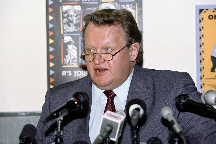 Martti Ahtisaari wearing glasses and a grey suit jacket in front of press microphones. 