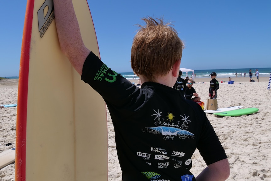 A boy with ginger hair leans on his surfboard.
