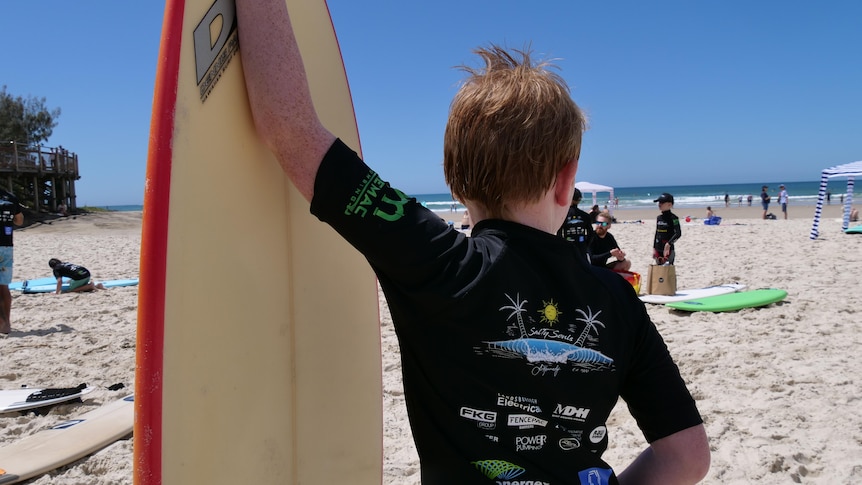 A boy with ginger hair leans on his surfboard.