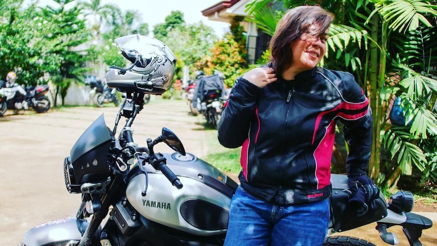 A woman in jeans and a black jacket leans against a motorcycle 