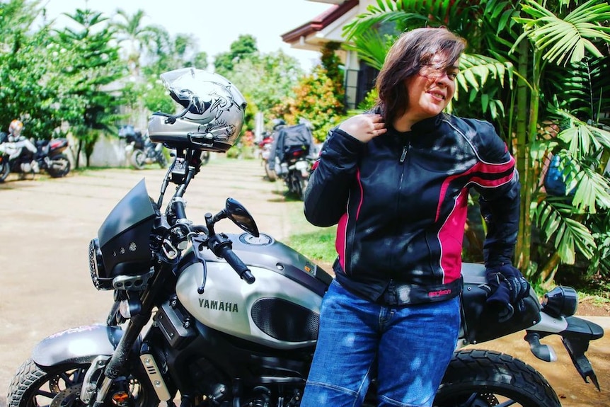 A woman in jeans and a black jacket leans against a motorcycle 