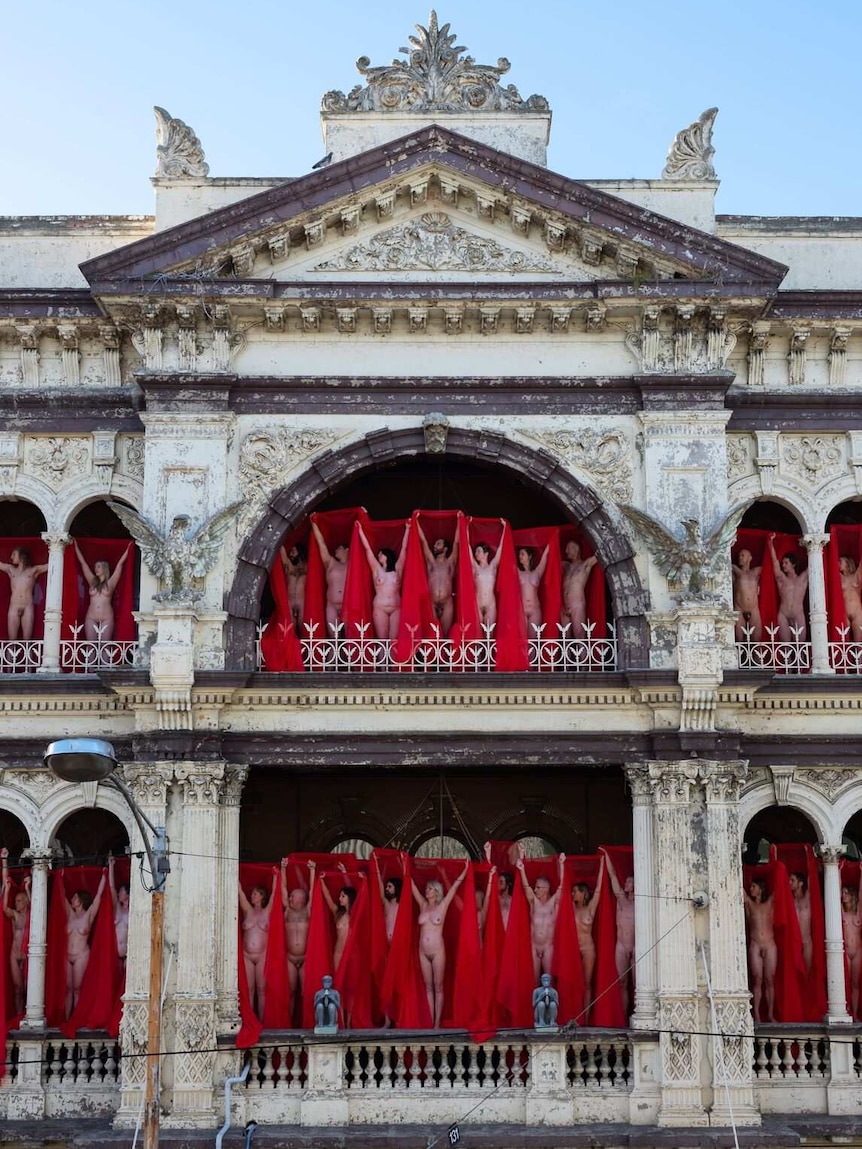 Naked people stand on balconies draped in red veils.