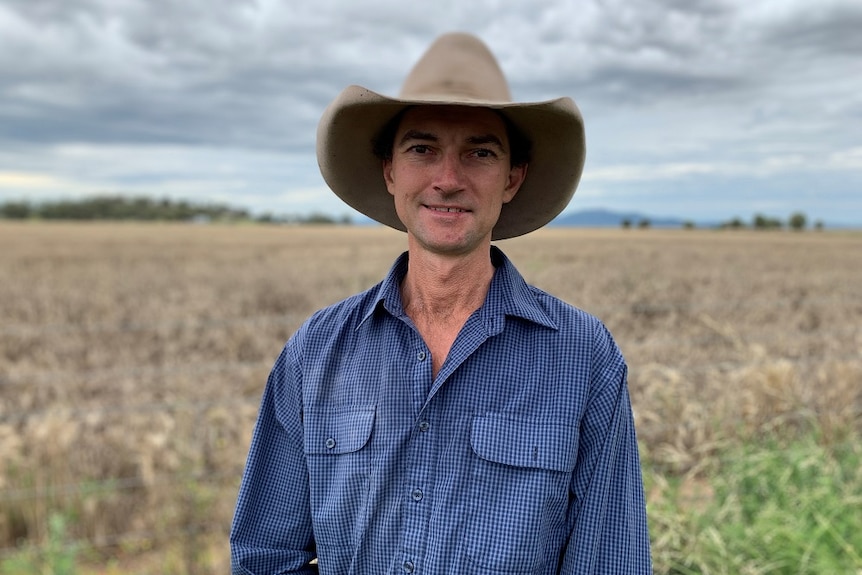 A farmer in a hat and work shirt stands in a paddock beneath a grey sky. 