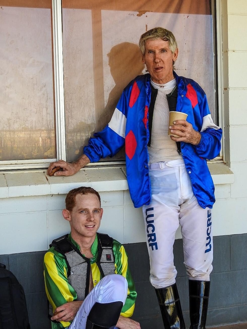 A jockey sits on the ground leaning up against a white brick wall next to a standing jockey