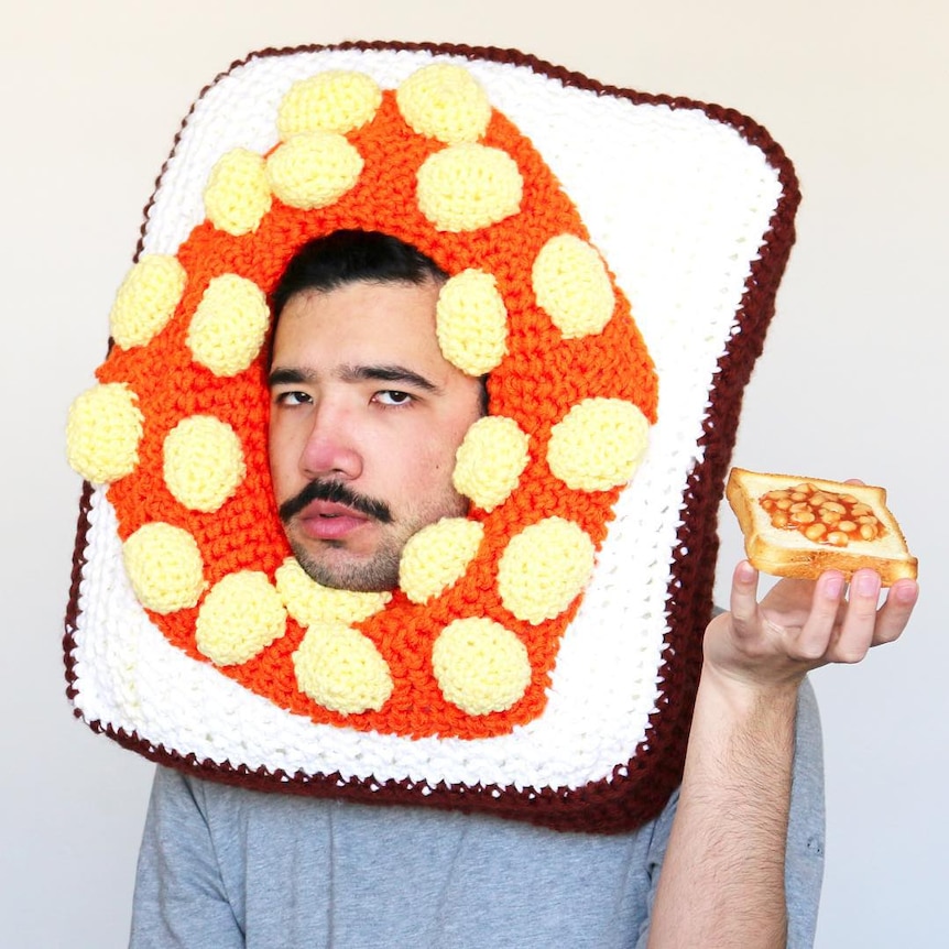 A man with a moustache holds up beans on toast, while wearing a crocheted headpiece in the shape of beans on toast