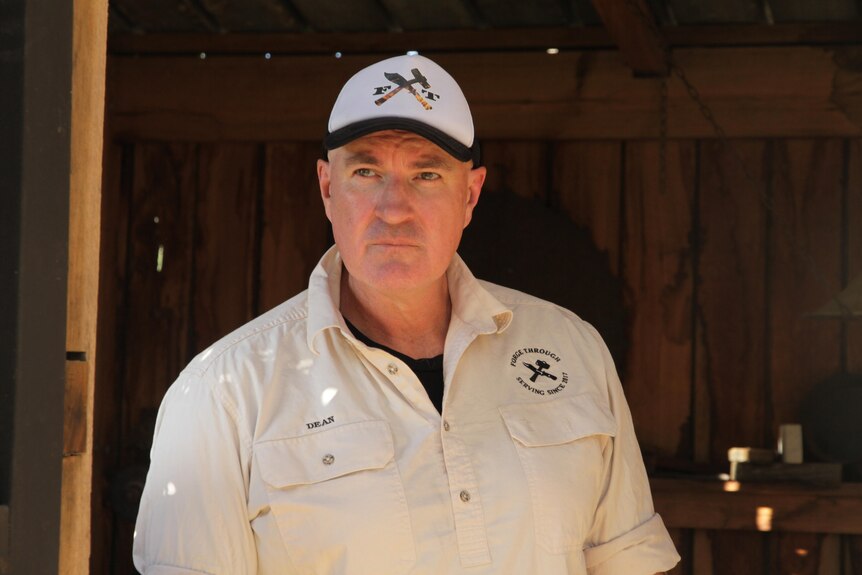 A serious man in a cream shirt and cap with a logo, a burning knife and hammer between F and T. He looks out into the distance.