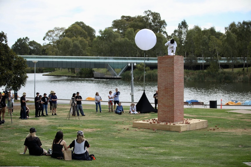 A crowd watches as someone performs atop a podium in a grass park alongside a river.