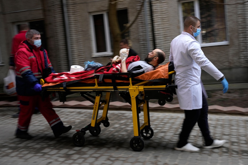 A man with bandaged hands lies on a wheeled stretcher, being pushed and pulled along by two other men in medical uniforms.