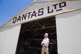A man in a white shirt stands outside an old airplane hangar with QANTAS LTD written it. An old propeller plane is behind him.