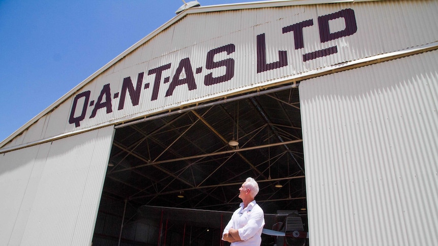 A man in a white shirt stands outside an old airplane hangar with QANTAS LTD written it. An old propeller plane is behind him.