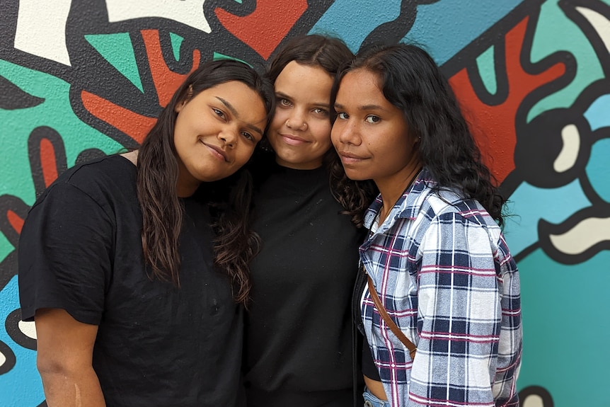 A picture of Janita Anderson, her sister Yatina and aunty Lizzy with in an embrace with subtle smiles in front of a mural