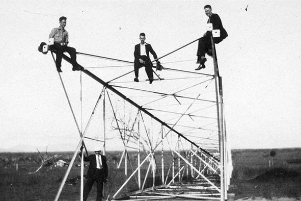 Three men sitting on top of a radio tower, one man standing underneath, black and white photo.