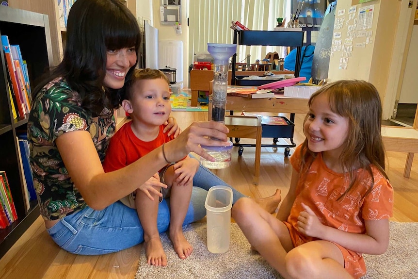A mother sits on a rug playing with a science experiment with a young boy and girl.