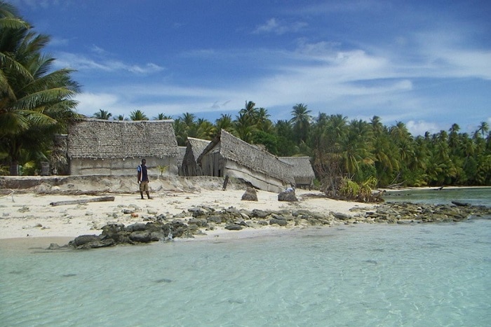Across clear Pacific waters, you see a group of huts with one sagging into the sea.