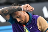 Tennis player Nick Kyrgios wipe brow with right arm, holding a tennis racquet in left hand.