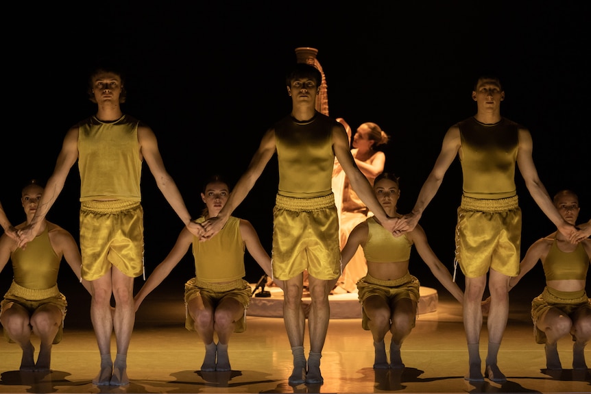 Stage shot showing  3 dancers in yellow tank tops and shorts standing and holding hands, with 4 crouching behind holding hands.