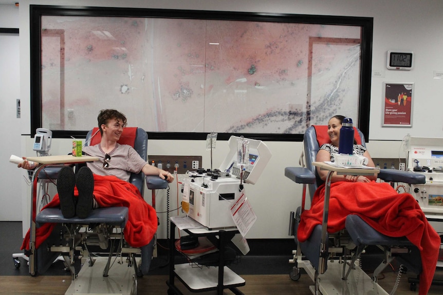 Two people sitting in blood donation chairs with machinery between them laugh while facing each other