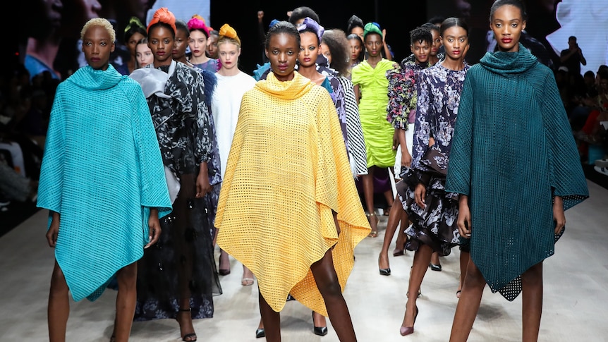 African women models pose on a runway, wearing an assortment of colourful garments.