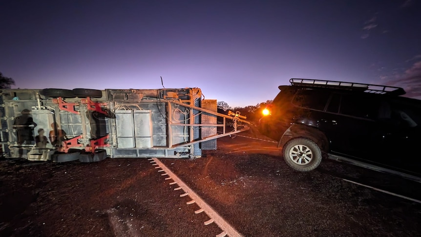 A campervan towed by a black 4WD lies on its side on a road at dusk.