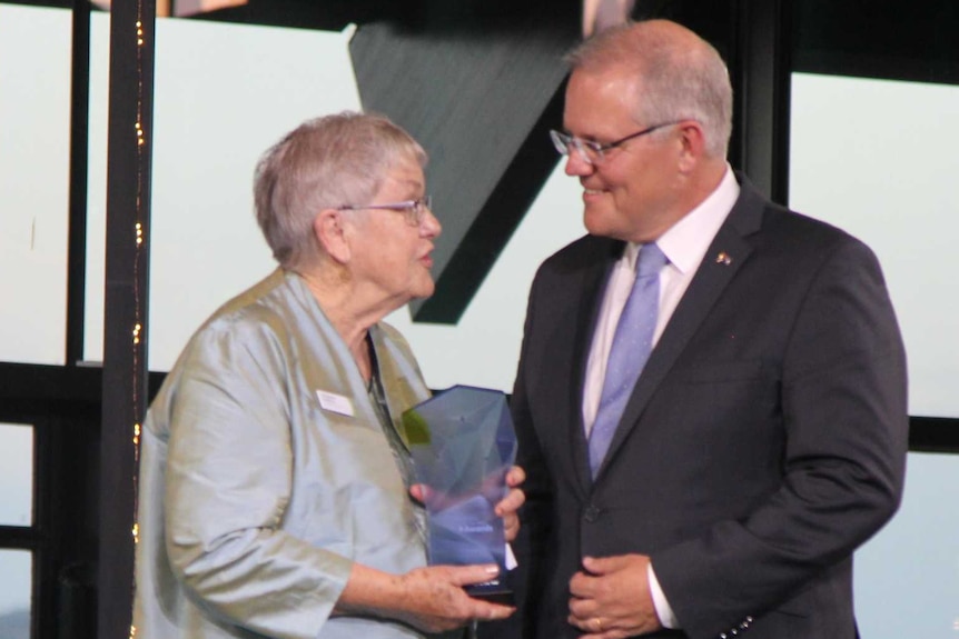 Sue Packer stands on stage holding the award for Senior Australian of the Year, talking to Prime Minister Scott Morrison.