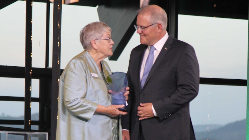Sue Packer stands on stage holding the award for Senior Australian of the Year, talking to Prime Minister Scott Morrison.