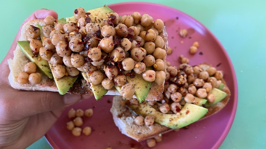 A pink plate is seen with two slices of toast on it, with sliced avocado and chickpeas on each. A hand holds one slice up.