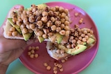 A pink plate is seen with two slices of toast on it, with sliced avocado and chickpeas on each. A hand holds one slice up.