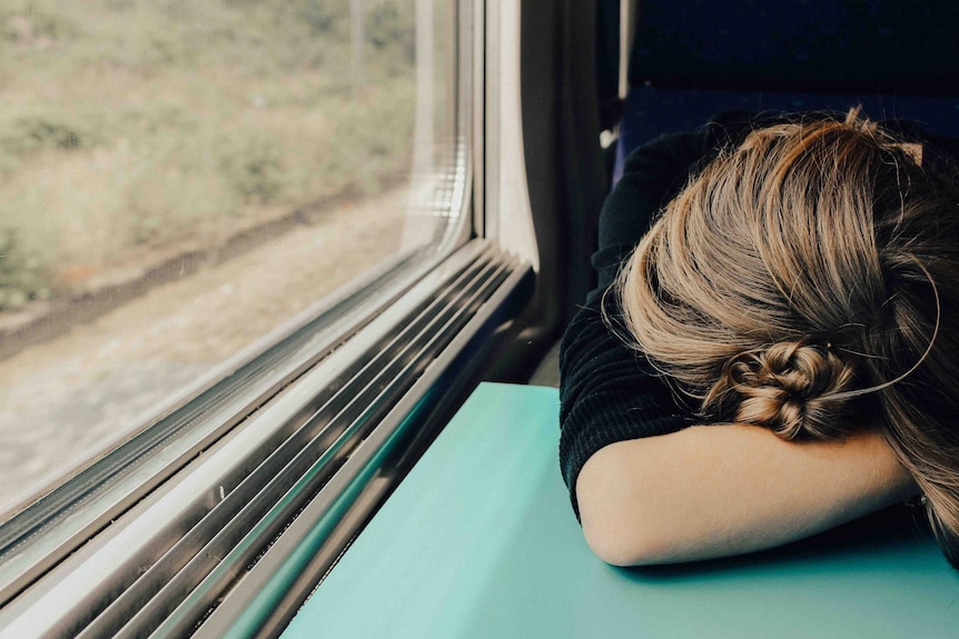 A woman asleep on a train rests her head against her forearm.